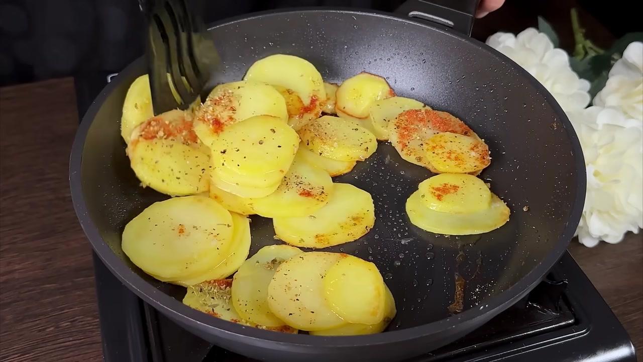 Cảnh quay cận cảnh chảo lớn với những lát khoai tây đang được chiên vàng giòn, một chiếc xẻng được dùng để đảo đều, phía sau là không gian bếp ấm cúng với lọ hoa trang trí.