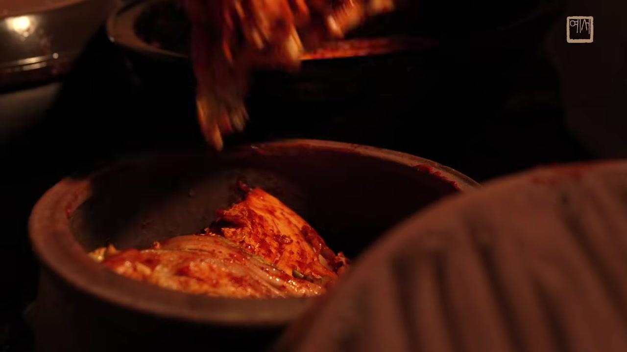Cảnh tượng những hũ đất sét lớn chứa đầy kimchi được đặt và lấp đất ở một góc vườn yên tĩnh, nơi có nền đất và đá.